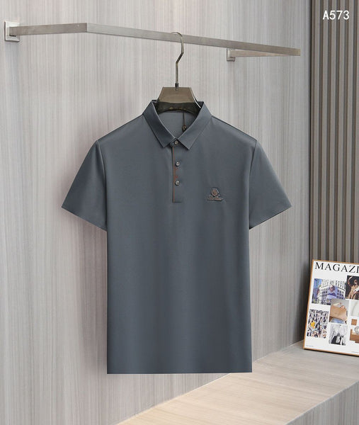 men's polo shirt hanging on a wooden hanger against a gray textured wall. The shirt features a classic spread collar, a partial button placket with decorative striped trim, and a small embroidered logo on the left chest. A magazine display is visible in the lower right corner of the background.