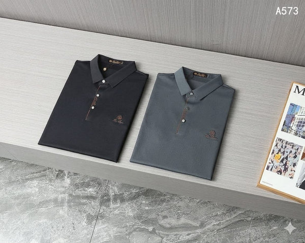 
men's polo shirt hanging on a wooden hanger against a gray textured wall. The shirt features a classic spread collar, a partial button placket with decorative striped trim, and a small embroidered logo on the left chest. A magazine display is visible in the lower right corner of the background.